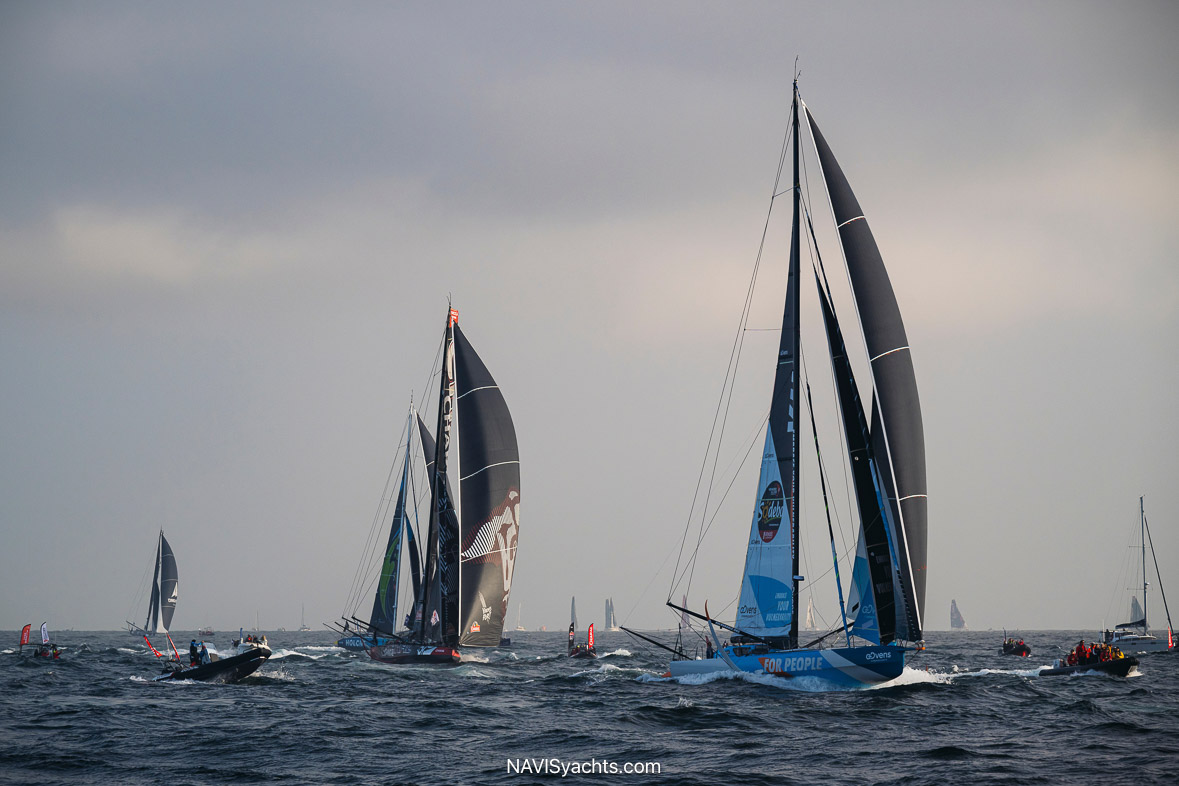 An IMOCA yacht with its foils extended, powerfully cutting through a large wave in the open ocean, showcasing the advanced technology and extreme conditions of the Vendée Globe race.