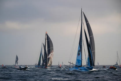 An IMOCA yacht with its foils extended, powerfully cutting through a large wave in the open ocean, showcasing the advanced technology and extreme conditions of the Vendée Globe race.